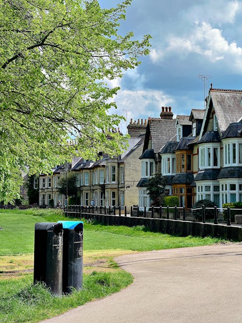 A row of terraced residential houses with bay windows and pitched roofs, situated behind a low black metal fence. In the foreground, a paved pathway runs alongside a grassy area where two black waste bins with blue lids are placed near a large leafy tree with fresh green foliage. The sky above is partly cloudy with patches of blue visible, suggesting a clear day. The scene appears to be in a suburban area, displaying a peaceful street environment suitable for local rubbish collection or private waste disposal services, with Waste Disposal Tufnell Park potentially facilitating rubbish removal from this residential setting.
