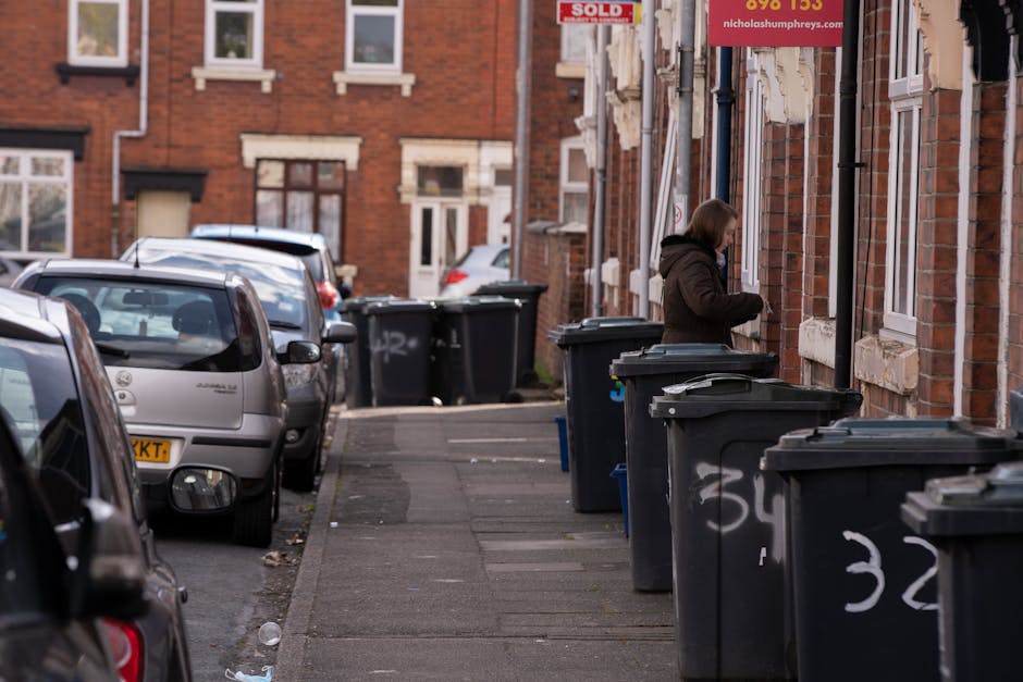 A row of black wheelie bins with closed lids lined up along the pavement outside a brick residential building in an urban street scene. The bins are positioned close to the building's exterior wall, which features multiple windows with white frames and stone sills. Several bins have white graffiti markings on their sides. A woman wearing a dark jacket with a hood stands next to one of the bins, appearing to handle or examine rubbish. In the background, additional bins are visible, placed along the sidewalk near similar brick houses with front doors and windows. Parked cars, including a silver hatchback and other vehicles, line the street to the left of the pavement, which is partially littered with small pieces of debris. The overall scene suggests an area where residents may conduct private waste disposal or rubbish collection services, with Waste Disposal Tufnell Park providing alternative waste handling options outside standard local authority services.