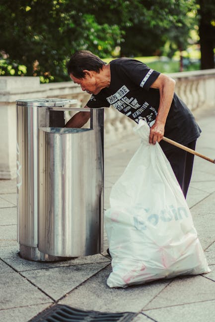 A middle-aged man with dark hair wearing a black T-shirt with white text leans forward to dispose of trash into a silver, cylindrical recycling bin with a slanted opening, situated on a paved sidewalk. He holds a white plastic bag filled with rubbish in his left hand, while his right arm rests on the bin's edge. The surface of the bin is metallic and reflective, showing subtle highlights and shadows. The background includes a stone balustrade and lush green trees, suggesting an outdoor urban environment. The lighting appears natural, indicative of a sunny day. This scene depicts independent waste disposal or rubbish removal, aligning with services offered by Waste Disposal Tufnell Park, situated in a setting appropriate for slip-on or on-site rubbish collection efforts.